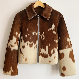 Könnte beinhalten: Eine braune und cremefarbene Kuhfelljacke mit Reißverschluss und Kragen. Die Jacke hat ein einzigartiges Muster und hängt an einem Holzbügel.