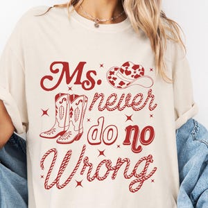 Könnte beinhalten: Cremefarbenes T-Shirt mit roter Grafik. Die Grafik enthält den Text "Ms. Never do no Wrong" mit Cowboyhut, Stiefeln und Sternen. Das Shirt hat kurze Ärmel und eine lockere Passform.