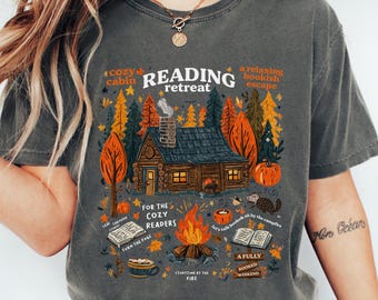 May include: Dark grey t-shirt with a colorful graphic of a cabin in a forest, a campfire, and the words "Reading Retreat." The design includes fall leaves, pumpkins, and text that reads "For the cozy readers" and "A fully booked weekend."