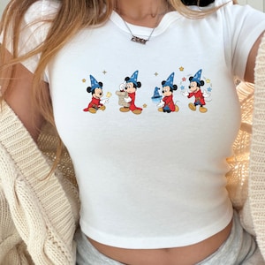 Puede incluir: Top corto blanco con un dise&ntilde;o de dibujos animados de Mickey Mouse con un disfraz de mago. El dise&ntilde;o muestra a Mickey en cuatro poses diferentes, cada una con un accesorio diferente. La camiseta tiene cuello redondo y mangas cortas.