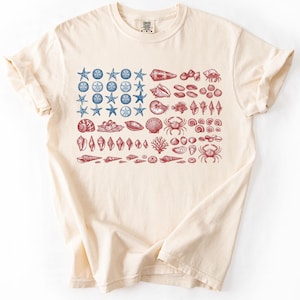 May include: Ivory-colored t-shirt featuring a design of various seashells and starfish in red and blue, arranged in a flag-like pattern. The shirt has a classic crew neck and short sleeves. The word "Ivory" is printed at the bottom left.
