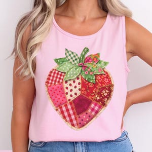 May include: Pink tank top featuring a large, patchwork strawberry design. The strawberry is composed of various fabric patterns in red, pink, and green, with a green and white checkered bow at the top. The word "Blossom" is in the bottom left corner.