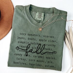 Op de afbeelding: Olijfgroen Comfort Colors T-shirt met het woord "fall" in cursief en een lijst met herfstthemawoorden. Een gouden ketting met een hanger is over het shirt gedrapeerd. Een bruine hoed is zichtbaar.