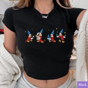 Puede incluir: Top corto negro con un gr&aacute;fico colorido de Mickey Mouse con un disfraz de mago. El dise&ntilde;o muestra a Mickey en diferentes poses con un sombrero de mago azul, una t&uacute;nica roja y estrellas. La camiseta es de manga corta.