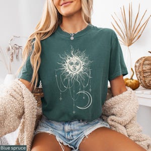 Puede incluir: Una camiseta de manga corta en color verde azulado oscuro con un estampado gr&aacute;fico blanco de un sol con rostro humano, una luna creciente y cadenas de cuentas colgantes con estrellas. La prenda tiene cuello redondo y corte holgado.