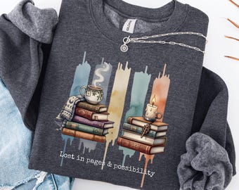 May include: Dark heather grey sweatshirt featuring a graphic of books, a teacup, and a candle. The text "Lost in pages & possibility" is printed below the graphic. A silver necklace with a sun pendant is draped across the sweatshirt.