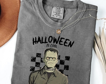 Puede incluir: Camiseta gris Comfort Colors con el texto "HALLOWEEN IS COOL" y un gráfico del monstruo de Frankenstein. La camiseta tiene cuello redondo y mangas cortas. Un collar plateado con un colgante está sobre la camiseta.