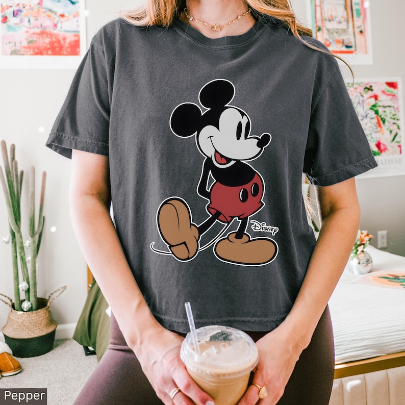 K&ouml;nnte beinhalten: Dunkelgraues T-Shirt mit einer gro&szlig;en Mickey Mouse-Grafik. Die Zeichentrickfigur ist in ihrer klassischen Pose dargestellt, mit schwarzen Ohren, einem wei&szlig;en Gesicht, roten Shorts und braunen Schuhen. Das Disney-Logo ist sichtbar.