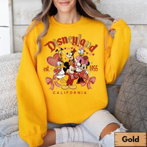 May include: A gold-coloured sweatshirt with a Disneyland graphic featuring Mickey Mouse, Minnie Mouse, Donald Duck, Goofy, and Daisy Duck. The graphic includes the text "Disneyland est. 1955 California". The sweatshirt has long sleeves and a crew neck.