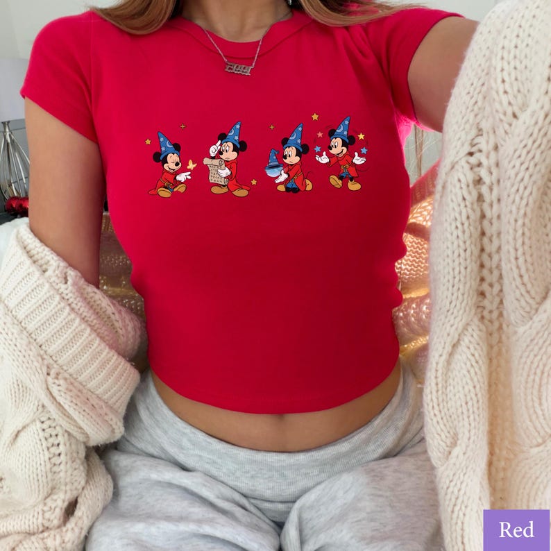 Puede incluir: Un top corto rojo con un gr&aacute;fico de dibujos animados de Mickey Mouse con un disfraz de mago. El gr&aacute;fico muestra a Mickey en cuatro poses diferentes. La camiseta tiene mangas cortas y cuello redondo. La palabra "Red" est&aacute; en un cuadro morado en la esquina inferior derecha.