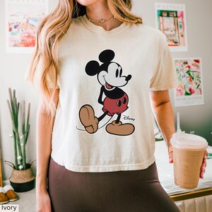 K&ouml;nnte beinhalten: Elfenbeinfarbenes T-Shirt mit Mickey Mouse-Grafik. Die Cartoonfigur ist in Schwarz, Wei&szlig;, Rot und Braun gehalten, mit dem Disney-Logo. Das T-Shirt hat eine lockere Passform. Das Wort "Ivory" ist unten links aufgedruckt.