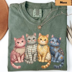 Può includere: T-shirt Comfort Colors verde muschio con quattro gatti dei cartoni animati con motivi e colori unici. I gatti sono rosa, grigi, arancioni e blu, ognuno con un design diverso. Una collana d'argento con un ciondolo è drappeggiata sulla maglietta.