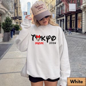 Pode incluir: Camisola branca com o texto "Tokyo Mom 2026" em preto e vermelho, com um gr&aacute;fico da Minnie Mouse. A camisola &eacute; combinada com cal&ccedil;&otilde;es pretos e um bon&eacute; de beisebol bege.
