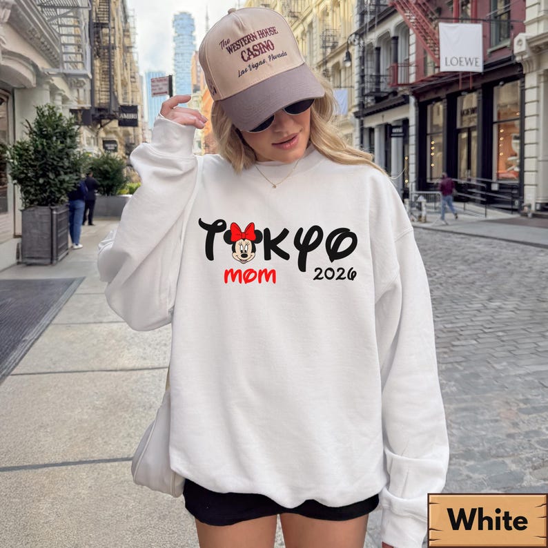Pode incluir: Camisola branca com o texto "Tokyo Mom 2026" em preto e vermelho, com um gr&aacute;fico da Minnie Mouse. A camisola &eacute; combinada com cal&ccedil;&otilde;es pretos e um bon&eacute; de beisebol bege.