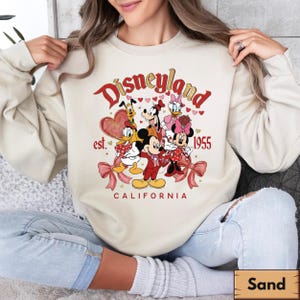 May include: A sand-coloured sweatshirt with a Disneyland graphic. The design features Mickey Mouse, Minnie Mouse, Donald Duck, Daisy Duck, Goofy, and Pluto. The text reads "Disneyland est. 1955 California" in red and gold.
