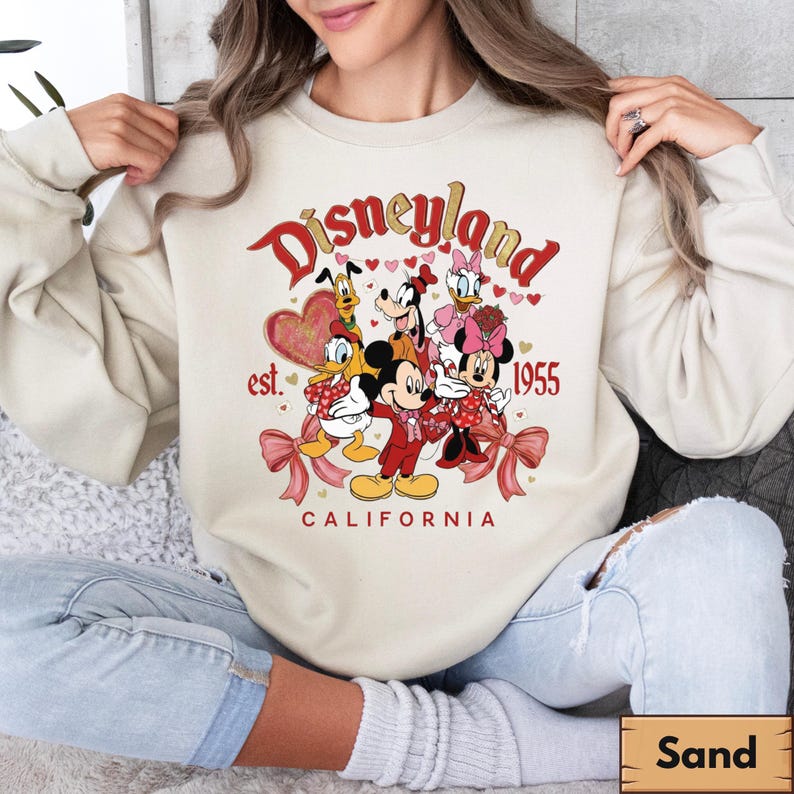 May include: A sand-coloured sweatshirt with a Disneyland graphic. The design features Mickey Mouse, Minnie Mouse, Donald Duck, Daisy Duck, Goofy, and Pluto. The text reads "Disneyland est. 1955 California" in red and gold.
