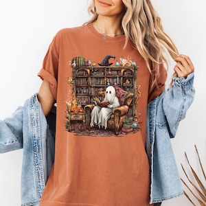 Könnte beinhalten: Gebrannt orangefarbenes T-Shirt mit einer Fantasy-Illustration. Es zeigt einen Geist, der in einem Sessel ein Buch liest, umgeben von einem Bücherregal voller Bücher, Tränke und einem Hexenhut. Das Design erinnert an Halloween oder Fantasy.