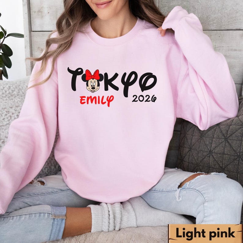 Pode incluir: Camisola rosa claro com a palavra "TOKYO" em preto, um gr&aacute;fico da Minnie Mouse, o nome "EMILY" em vermelho e o ano "2026" em preto. A camisola &eacute; de cor rosa claro lisa.