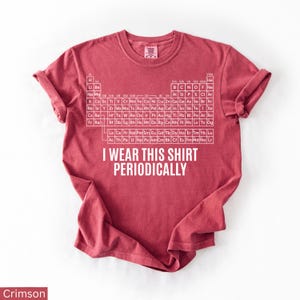 May include: Crimson-colored t-shirt featuring the periodic table of elements in white. The shirt includes the text "I WEAR THIS SHIRT PERIODICALLY" in white. The shirt is a crew neck and has rolled-up sleeves.