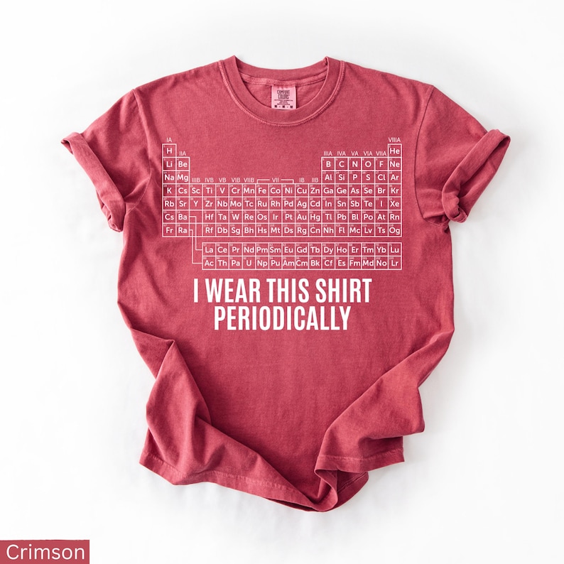 May include: Crimson-colored t-shirt featuring the periodic table of elements in white. The shirt includes the text "I WEAR THIS SHIRT PERIODICALLY" in white. The shirt is a crew neck and has rolled-up sleeves.
