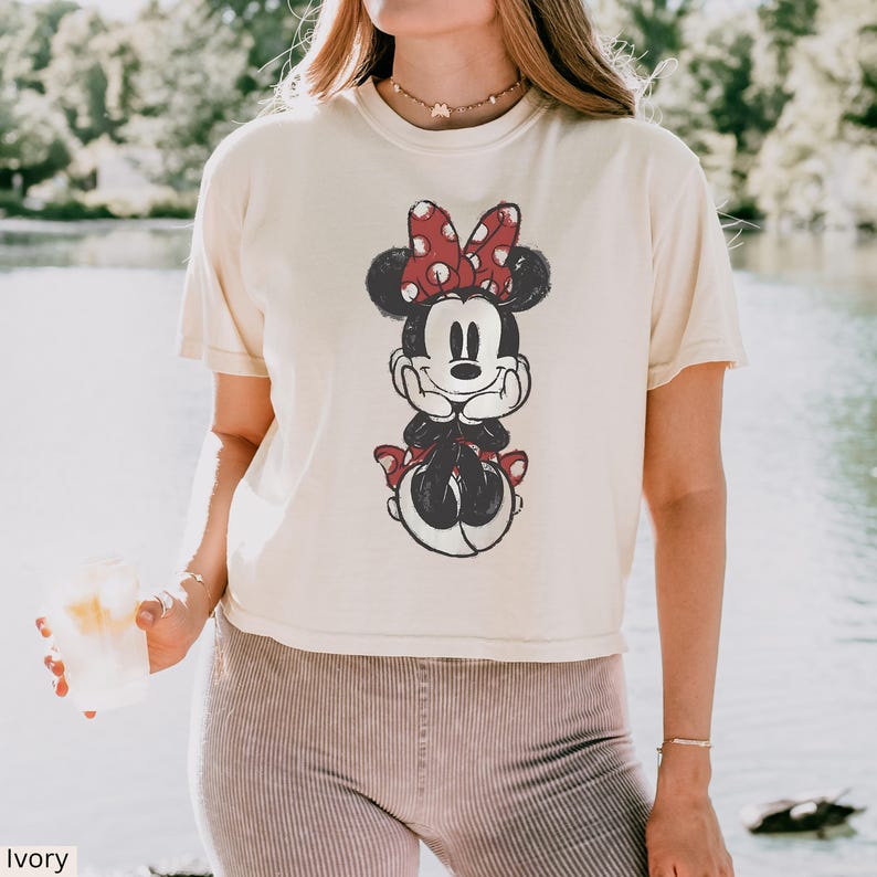 May include: Ivory t-shirt with a distressed Minnie Mouse graphic. The design shows Minnie with a red polka-dot bow, black ears, and a smiling expression. The top has short sleeves and a relaxed fit, suitable for everyday wear.