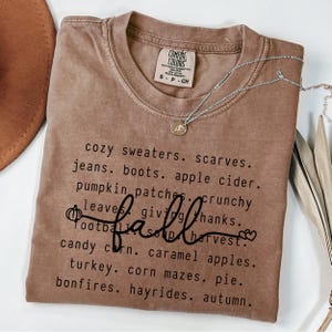 Op de afbeelding: Een bruin Comfort Colors t-shirt met het woord "fall" in cursief en een lijst met herfstthemawoorden. Een zilveren ketting met een hanger is over het shirt gedrapeerd.