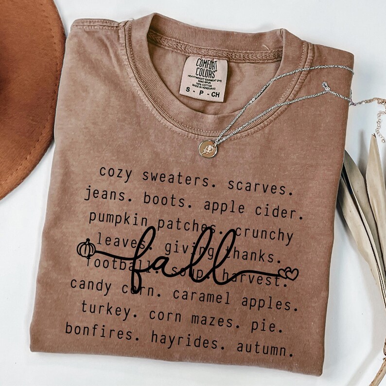 Op de afbeelding: Een bruin Comfort Colors t-shirt met het woord "fall" in cursief en een lijst met herfstthemawoorden. Een zilveren ketting met een hanger is over het shirt gedrapeerd.
