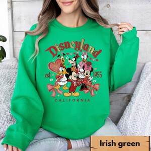 May include: Green sweatshirt with a Disneyland graphic featuring Mickey Mouse, Minnie Mouse, Donald Duck, Daisy Duck, Goofy, and Pluto. The graphic includes the text "Disneyland est. 1955 California" in red and white.