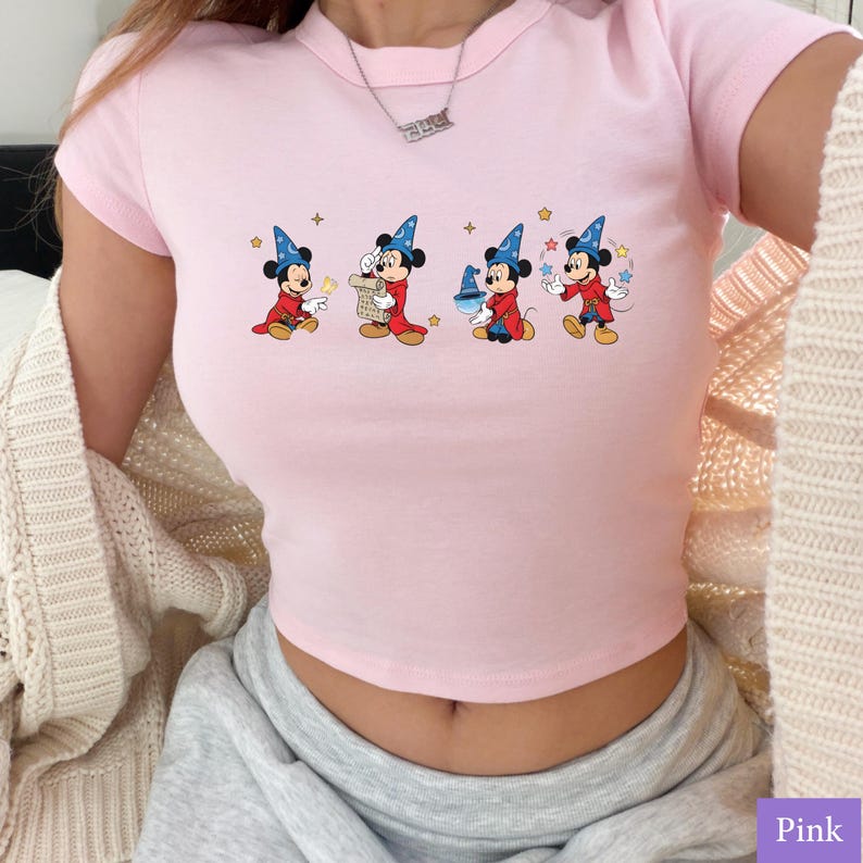 Puede incluir: Top corto rosa claro con un dise&ntilde;o de dibujos animados de Mickey Mouse vestido de mago. El dise&ntilde;o muestra a Mickey en cuatro poses diferentes, cada una con un sombrero y una t&uacute;nica de mago. La camiseta tiene cuello redondo y mangas cortas.