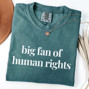 Pode incluir: Uma camiseta Comfort Colors verde-azulado com a frase "big fan of human rights" em letras brancas. Um colar de ouro com um pingente repousa sobre a camisa. Um chapéu marrom e caules de plantas secas estão no fundo.