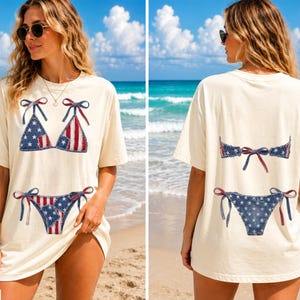 Pode incluir: Camiseta de cor marfim com um design de biquíni patriótico. A frente mostra um top e uma parte de baixo de biquíni com um padrão de bandeira americana vermelho, branco e azul. A parte de trás da camisa também mostra o design do biquíni. Perfeito para um dia de praia de verão.