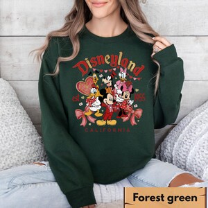 May include: A forest green sweatshirt with a Disneyland graphic. The design includes Mickey Mouse, Minnie Mouse, Donald Duck, and other Disney characters. The text reads "Disneyland California est. 1955".