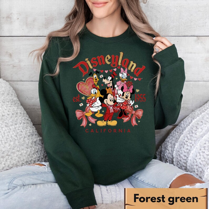 May include: A forest green sweatshirt with a Disneyland graphic. The design includes Mickey Mouse, Minnie Mouse, Donald Duck, and other Disney characters. The text reads "Disneyland California est. 1955".