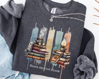 May include: Dark heather grey sweatshirt with a graphic of books, a coffee cup, and a lantern. The design includes the text "Where stories bloom" and watercolour-style accents. A silver necklace is draped across the top of the sweatshirt.