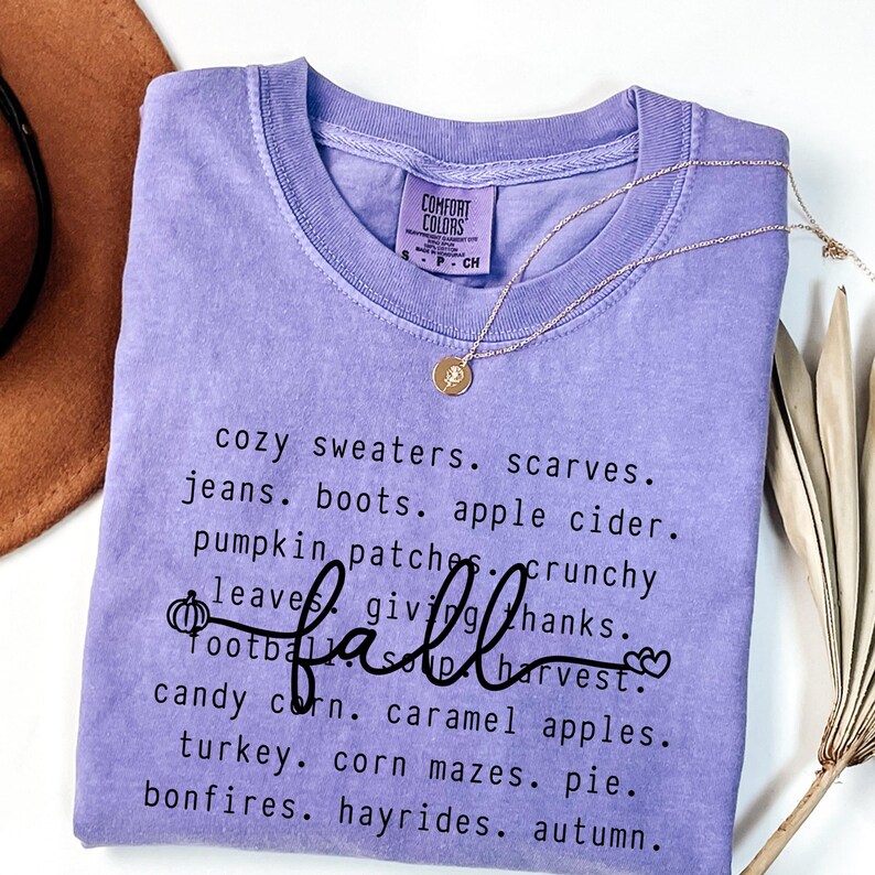 Op de afbeelding: Een lichtpaars Comfort Colors T-shirt met het woord "fall" in cursief en een lijst met herfstthemawoorden. Een gouden ketting met een hanger en een bruine hoed zijn ook zichtbaar.