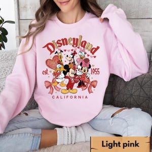 May include: Pale pink sweatshirt with a Disneyland graphic featuring Mickey and Minnie Mouse, Donald Duck, Goofy, Pluto, and Daisy Duck. The graphic includes the text "Disneyland est. 1955 California". The sweatshirt has long sleeves.