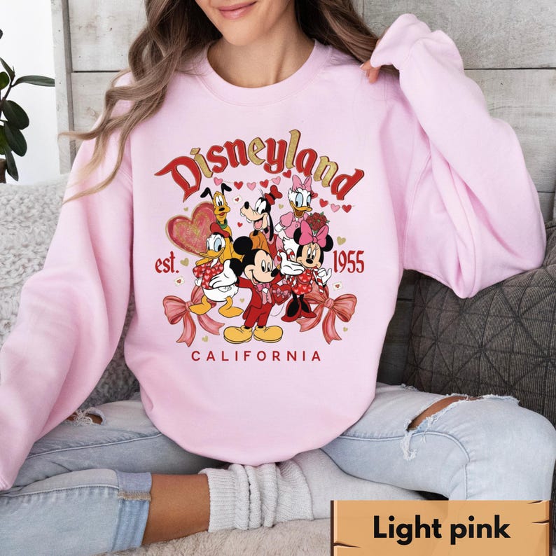May include: Pale pink sweatshirt with a Disneyland graphic featuring Mickey and Minnie Mouse, Donald Duck, Goofy, Pluto, and Daisy Duck. The graphic includes the text "Disneyland est. 1955 California". The sweatshirt has long sleeves.