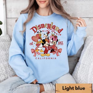 May include: Pale blue sweatshirt with a Disneyland graphic. The design features Mickey and Minnie Mouse, Donald and Daisy Duck, and Goofy. The text reads "Disneyland est. 1955 California" in red and gold. The graphic includes hearts and bows.