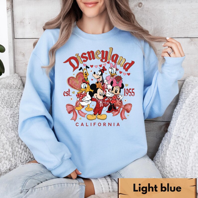 May include: Pale blue sweatshirt with a Disneyland graphic. The design features Mickey and Minnie Mouse, Donald and Daisy Duck, and Goofy. The text reads "Disneyland est. 1955 California" in red and gold. The graphic includes hearts and bows.