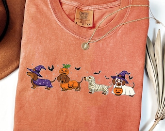 May include: A coral-colored t-shirt featuring a Halloween-themed dachshund design. The graphic includes four dachshunds dressed as a witch, a pumpkin, a mummy, and a ghost, with bats. The shirt has a Comfort Colors tag and a gold necklace.