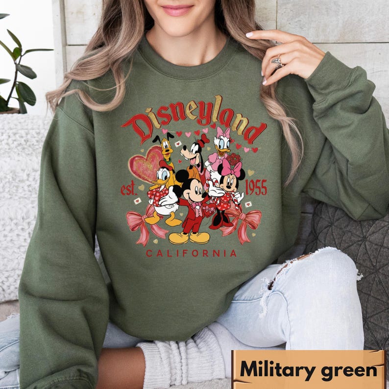 May include: Military green sweatshirt with a Disneyland graphic. The design features Mickey Mouse, Minnie Mouse, Donald Duck, Daisy Duck, Goofy, and Pluto. The text reads "Disneyland California est. 1955" in red and gold.