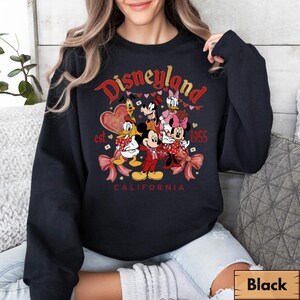 May include: Black sweatshirt with a Disneyland graphic featuring Mickey Mouse, Minnie Mouse, Donald Duck, Daisy Duck, Goofy, and Pluto. The graphic includes the text "Disneyland California est. 1955" in red and gold.