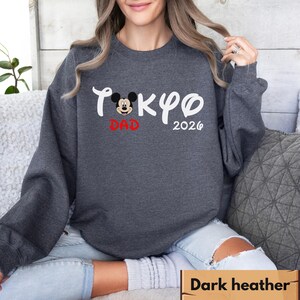 Pode incluir: Camisola cinza escuro com o texto "Tokyo Dad 2026" em escrita branca e um gr&aacute;fico do Mickey Mouse. A palavra "Dad" &eacute; vermelha. A camisola &eacute; de gola redonda.