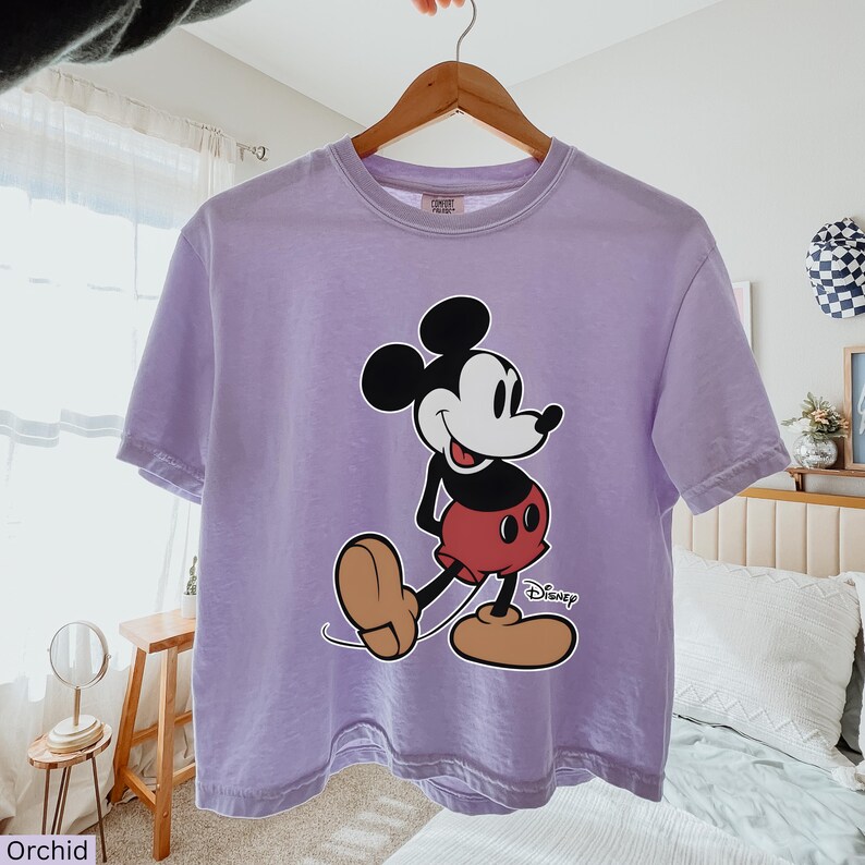 K&ouml;nnte beinhalten: Ein helllila T-Shirt mit einer Mickey Mouse-Grafik. Die Zeichentrickfigur ist in Schwarz, Rot und Braun dargestellt. Das T-Shirt h&auml;ngt an einem Holzb&uuml;gel.