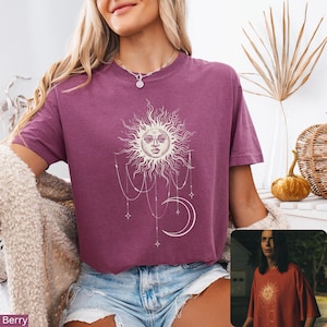 Puede incluir: Una camiseta de manga corta color baya con un estampado gr&aacute;fico blanco de un sol con cara, una luna creciente y cadenas de cuentas colgantes con estrellas. La prenda tiene cuello redondo y se combina con pantalones cortos de mezclilla.