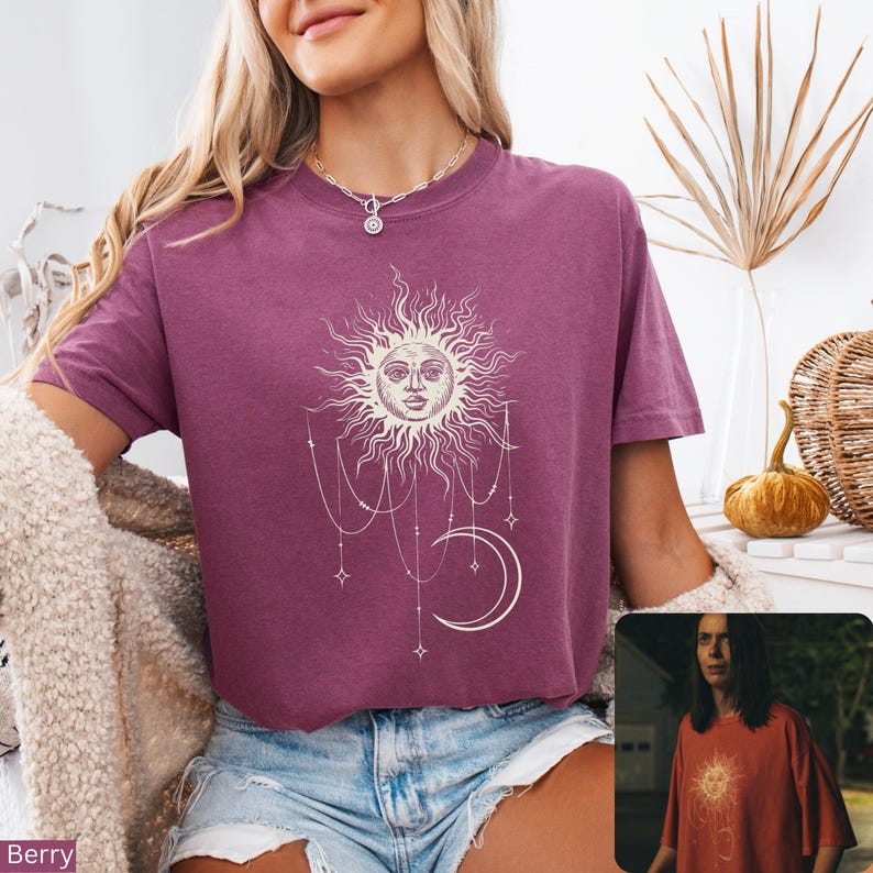 Puede incluir: Una camiseta de manga corta color baya con un estampado gr&aacute;fico blanco de un sol con cara, una luna creciente y cadenas de cuentas colgantes con estrellas. La prenda tiene cuello redondo y se combina con pantalones cortos de mezclilla.