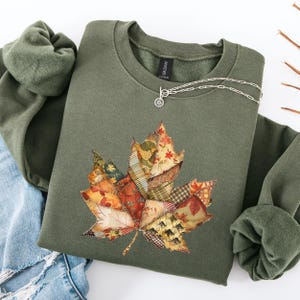 May include: An olive green sweatshirt with a large, patchwork-style maple leaf design in autumn colors. A silver necklace with a small pendant is draped on the front. The sweatshirt is folded with a portion of a blue jean visible.