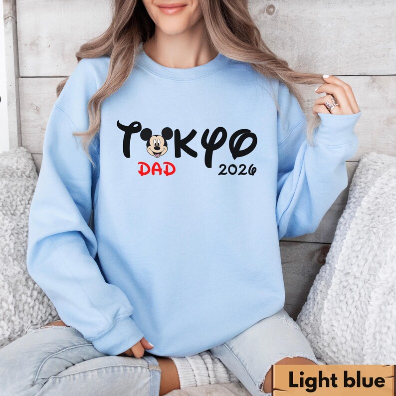 Pode incluir: Camisola azul clara com o texto "Tokyo Dad 2026" em preto e vermelho. A palavra "Tokyo" apresenta uma cabe&ccedil;a do Mickey Mouse no lugar do "o". A camisola &eacute; de cor azul clara lisa.