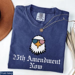 May include: True navy blue t-shirt with a graphic of an eagle wearing sunglasses with an American flag design. The text "25th Amendment Now" is printed in white script below the eagle. A gold necklace is draped across the shirt.