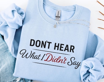 May include: Light blue sweatshirt with the text "DON'T HEAR What I Didn't Say" in black and red. A silver necklace with a small pendant is on the sweatshirt. The sweatshirt is folded on top of a pair of blue jeans.